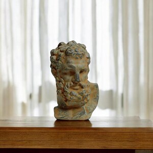 May include: A weathered gray and brown stone bust of a man with a beard. The bust is on a wooden surface.