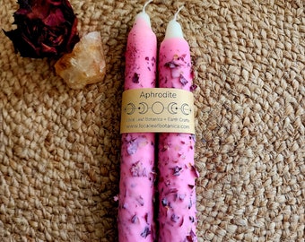 May include: Two pink candles with a white top and adorned with dried rose petals. The candles are labeled "Aphrodite" with a brown band. The candles are on a woven, brown surface.