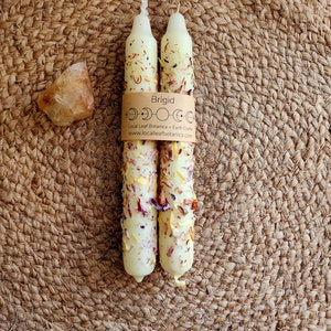 Könnte beinhalten: Zwei cremefarbene Kerzen mit floralen und Kräuterakzenten sind mit einem braunen Papierband gebündelt, auf dem "Brigid" steht. Ein kleiner, grob geschnittener, bernsteinfarbener Kristall liegt links von den Kerzen. Der Hintergrund ist eine gewebte, kreisförmige Matte.