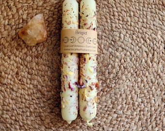 May include: Two cream-colored candles with floral and herb accents are bundled together with a brown paper band that reads "Brigid." A small, rough-cut, amber-colored crystal sits to the left of the candles. The background is a woven, circular mat.