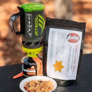 May include: A camping breakfast setup featuring a Jetboil stove and a bag of Mamoo's Camp Kitchen breakfast scramble. A small bowl of prepared scramble is in the foreground. The bag's label describes the ingredients: eggs, cheese, sausage, and potatoes.