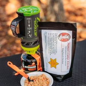 May include: A camping meal setup featuring a Jetboil stove, a pouch of King Ranch Chicken, and a bowl of prepared food. The stove is black, gray, and green, with an orange fuel canister. The food is served with an orange utensil.
