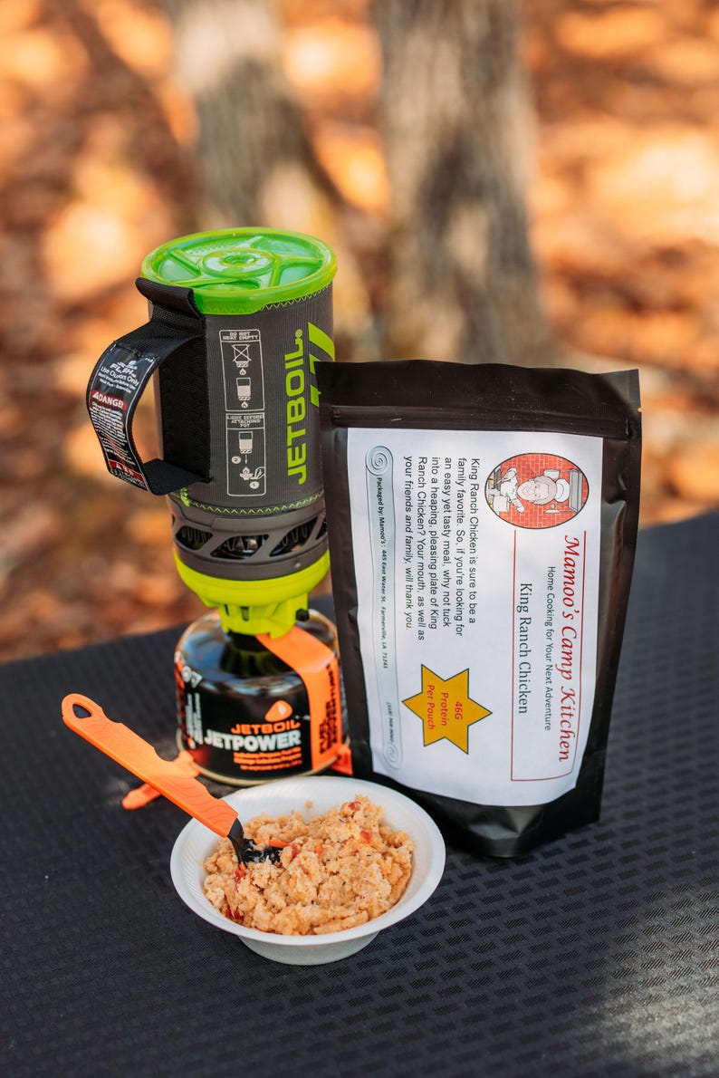 May include: A camping meal setup featuring a Jetboil stove, a pouch of King Ranch Chicken, and a bowl of prepared food. The stove is black, gray, and green, with an orange fuel canister. The food is served with an orange utensil.