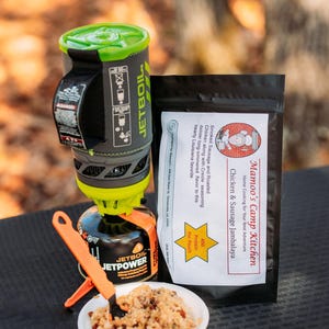 May include: A portable camping stove with a green and gray design, a black fuel canister, and a white bowl of food. A black pouch of "Mamoo's Camp Kitchen" Chicken & Sausage Jambalaya is also present. Orange utensils are included.