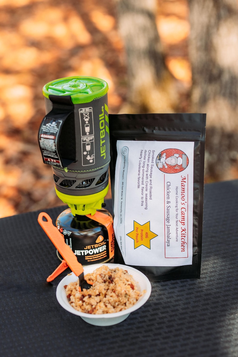 May include: A portable camping stove with a green and gray design, a black fuel canister, and a white bowl of food. A black pouch of "Mamoo's Camp Kitchen" Chicken & Sausage Jambalaya is also present. Orange utensils are included.