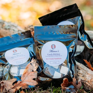 May include: Three sealed bags of ice cream sandwiches from Mamoo's Candy Kitchen are displayed on a bed of fallen leaves and weathered wood. Each bag has a white label with the brand's logo and product information. The background is blurred with natural light.