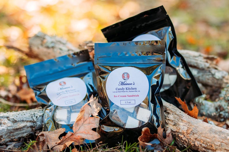 May include: Three sealed bags of ice cream sandwiches from Mamoo's Candy Kitchen are displayed on a bed of fallen leaves and weathered wood. Each bag has a white label with the brand's logo and product information. The background is blurred with natural light.