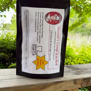 May include: A black pouch of Mamoo's Camp Kitchen Chicken & Sausage Gumbo. The label features text including "Smoked Sausage and Roasted Chicken" and "32G Protein Per Pouch." The product is certified Cajun and packaged in Farmerville, LA.