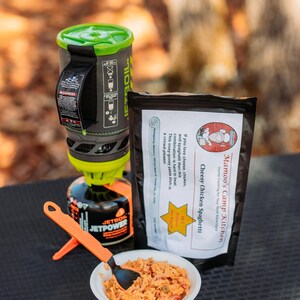 May include: A camping meal setup featuring a Jetboil stove, a pouch of Cheesy Chicken Spaghetti, and a bowl of prepared food. The stove is black, green, and orange. The food is in a white bowl with an orange spoon.