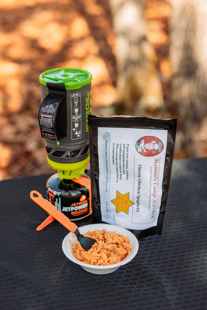 May include: A camping meal setup featuring a Jetboil stove, a pouch of Cheesy Chicken Spaghetti, and a bowl of prepared food. The stove is black, green, and orange. The food is in a white bowl with an orange spoon.