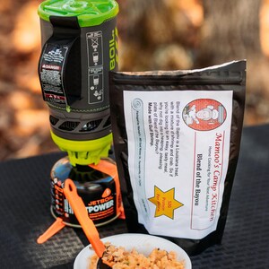 May include: A camping meal setup featuring a Jetboil stove, a bag of "Mamao's Camp Kitchen Blend of the Bayou", and a bowl of prepared food. The meal includes shrimp and crab. An orange spoon rests in the bowl. The stove is black and green.