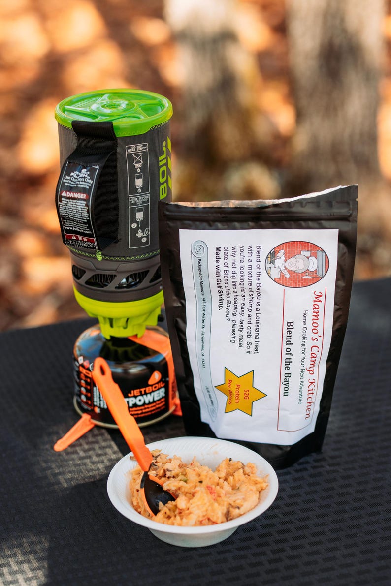 May include: A camping meal setup featuring a Jetboil stove, a bag of "Mamao's Camp Kitchen Blend of the Bayou", and a bowl of prepared food. The meal includes shrimp and crab. An orange spoon rests in the bowl. The stove is black and green.