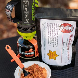May include: A camping meal setup featuring a Jetboil cooking system, a pouch of "Mamoo's Camp Kitchen Beefy Enchilada", and a bowl of prepared food with an orange spoon. The meal is ready for a camping adventure.
