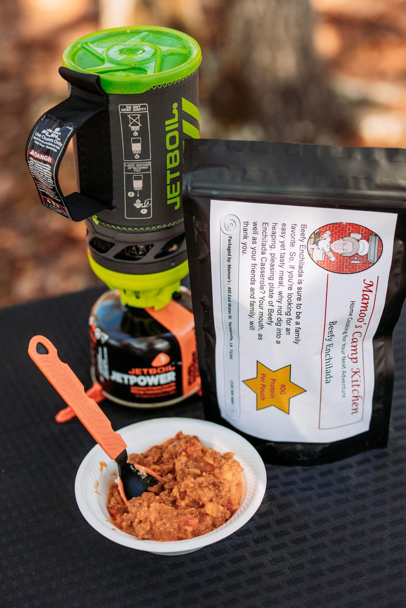 May include: A camping meal setup featuring a Jetboil cooking system, a pouch of "Mamoo's Camp Kitchen Beefy Enchilada", and a bowl of prepared food with an orange spoon. The meal is ready for a camping adventure.