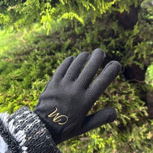 Peut inclure: Gants en tricot noir avec un "CM" brodé en or sur le dos de la main.