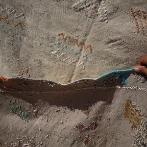 May include: Beige woven rug with colourful embroidered patterns. The rug features geometric and abstract designs in shades of orange, blue, and white. The edges are frayed, giving it a rustic appearance. The rug is made of a textured material.
