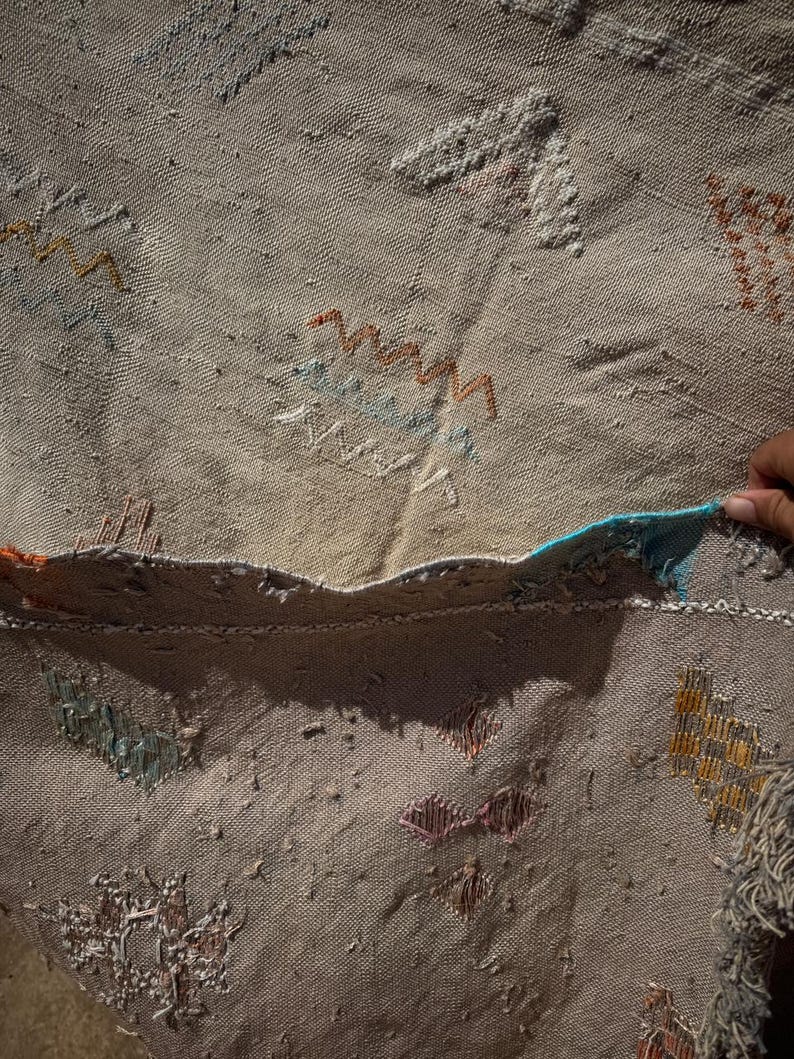 May include: Beige woven rug with colourful embroidered patterns. The rug features geometric and abstract designs in shades of orange, blue, and white. The edges are frayed, giving it a rustic appearance. The rug is made of a textured material.