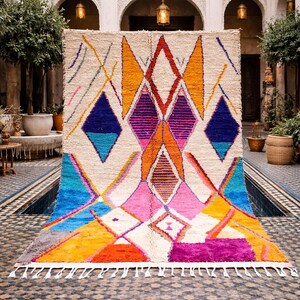 May include: A vibrant, geometric rug with a cream-colored background and a colorful diamond pattern. The design incorporates shades of blue, orange, pink, purple, and yellow. The rug has a fringe along the bottom edge and is displayed in an outdoor setting.