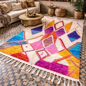 May include: A colorful, geometric patterned rug with a cream-colored background. The rug features a variety of shapes in shades of blue, pink, orange, yellow, and purple. The rug has a fringe along the bottom edge. This rug is perfect for a living room.