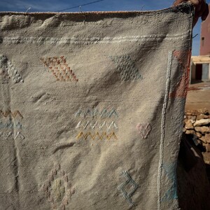 May include: Beige woven textile with colourful geometric embroidery. The textile features diamond, triangle, and zigzag patterns in shades of orange, blue, yellow, and pink. The edges are finished with a white border.