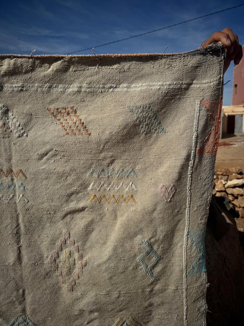 May include: Beige woven textile with colourful geometric embroidery. The textile features diamond, triangle, and zigzag patterns in shades of orange, blue, yellow, and pink. The edges are finished with a white border.