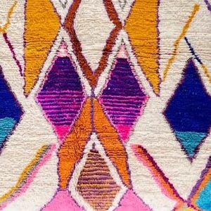 May include: Close-up of a colorful, textured rug with a geometric pattern. The design features diamond shapes in shades of orange, pink, purple, blue, and yellow against a white background. The rug has a thick, plush texture.