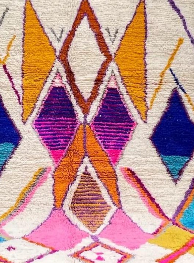 May include: Close-up of a colorful, textured rug with a geometric pattern. The design features diamond shapes in shades of orange, pink, purple, blue, and yellow against a white background. The rug has a thick, plush texture.