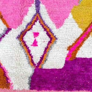 May include: Close-up of a colorful rug with a geometric pattern. The rug features a combination of pink, white, yellow, orange, and purple colors. The design includes diamond and triangle shapes.