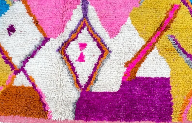 May include: Close-up of a colorful rug with a geometric pattern. The rug features a combination of pink, white, yellow, orange, and purple colors. The design includes diamond and triangle shapes.