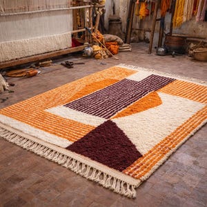 May include: A rectangular rug with a geometric design in shades of orange, cream, and burgundy. The rug has a textured surface and a fringe along the edges. The rug is displayed on a tiled floor in a workshop setting.