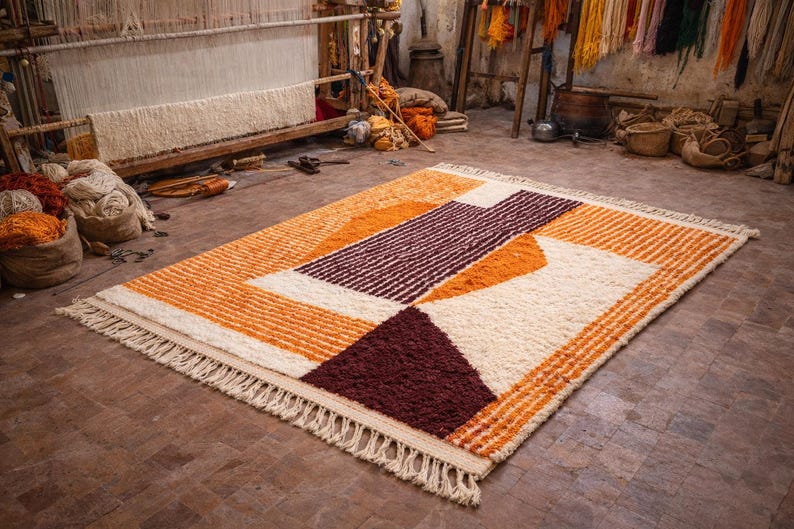 May include: A rectangular rug with a geometric design in shades of orange, cream, and burgundy. The rug has a textured surface and a fringe along the edges. The rug is displayed on a tiled floor in a workshop setting.