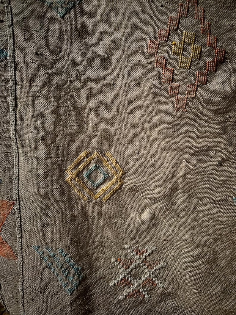 May include: Close-up of a woven textile with a brown background. The textile features geometric patterns in shades of yellow, orange, blue, and white. The patterns include diamond and star shapes, adding visual interest to the fabric. The texture of the fabric is visible.