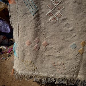 May include: Close-up of a beige woven rug with colourful geometric patterns. The rug features diamond and star-like designs in shades of blue, orange, pink, and yellow. The bottom edge has a fringe.