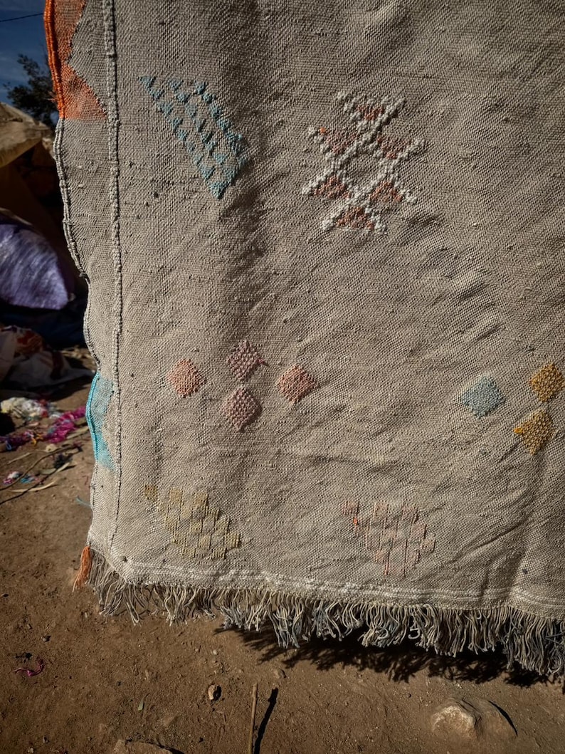 May include: Close-up of a beige woven rug with colourful geometric patterns. The rug features diamond and star-like designs in shades of blue, orange, pink, and yellow. The bottom edge has a fringe.
