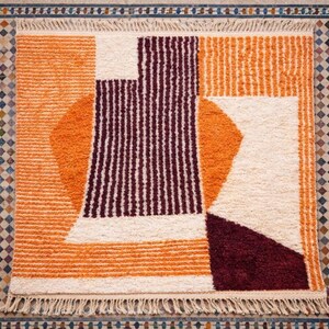 May include: A geometric patterned rug with a cream, orange, and burgundy color scheme. The rug features a combination of shapes, including rectangles, circles, and stripes. The rug has a fringed edge.
