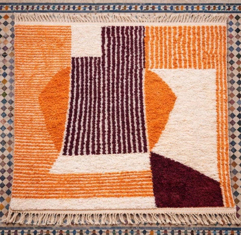 May include: A geometric patterned rug with a cream, orange, and burgundy color scheme. The rug features a combination of shapes, including rectangles, circles, and stripes. The rug has a fringed edge.