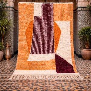 May include: A geometric patterned rug with a cream fringe. The rug features a design of orange, cream, and burgundy shapes. The rug is displayed in an outdoor setting with an arched doorway and potted plants.