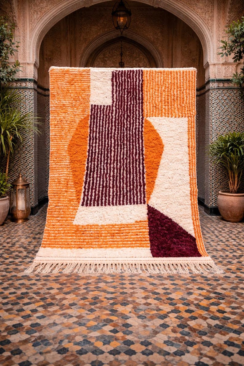 May include: A geometric patterned rug with a cream fringe. The rug features a design of orange, cream, and burgundy shapes. The rug is displayed in an outdoor setting with an arched doorway and potted plants.