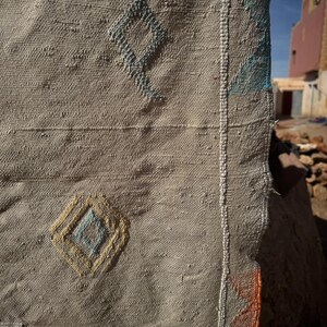May include: Close-up of a beige woven textile with diamond patterns in blue, yellow, and brown. The textile has a textured surface and a visible seam with white stitching. The background shows a blurred view of buildings.