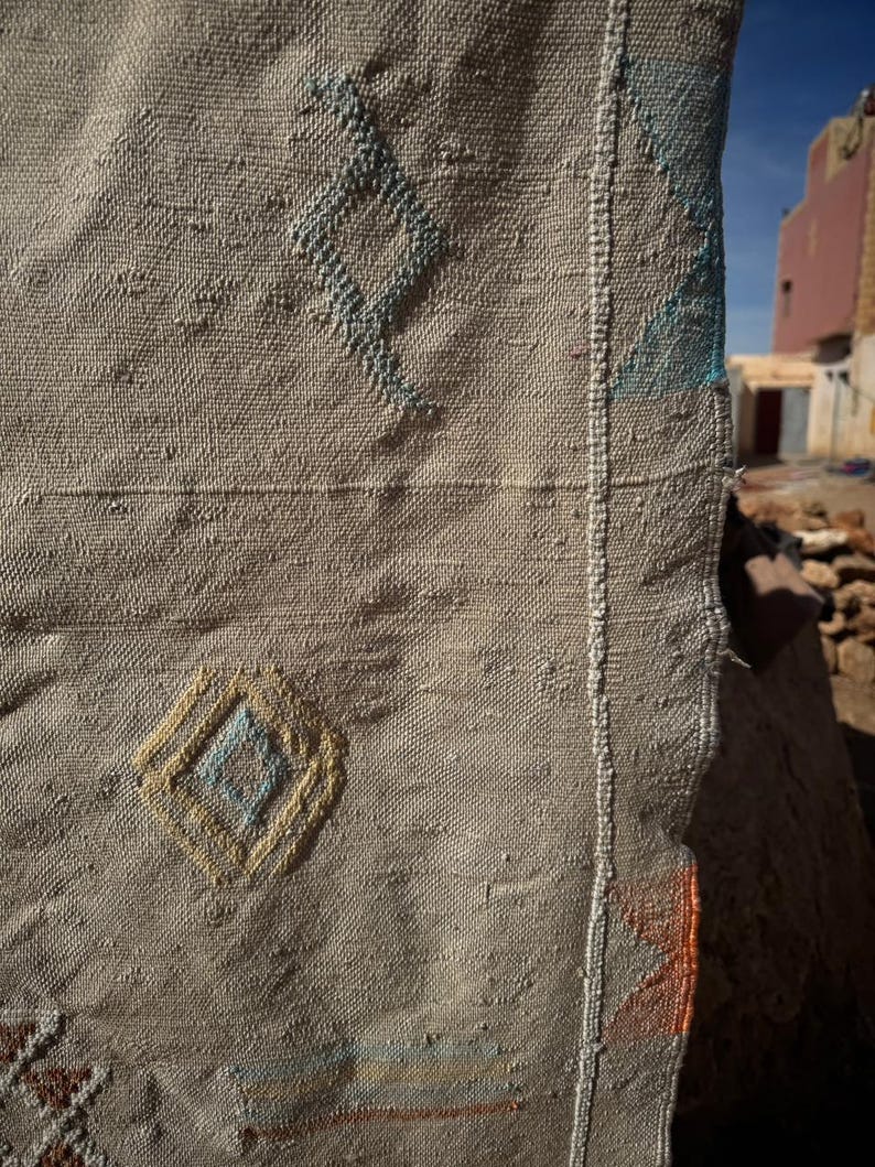 May include: Close-up of a beige woven textile with diamond patterns in blue, yellow, and brown. The textile has a textured surface and a visible seam with white stitching. The background shows a blurred view of buildings.