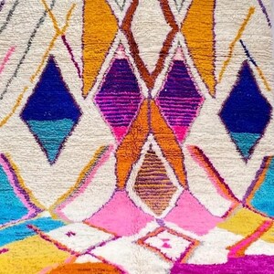 May include: Close-up of a colorful, geometric rug with a cream-colored background. The rug features diamond and triangle shapes in shades of blue, pink, orange, yellow, and purple. The rug has a textured, woven appearance.
