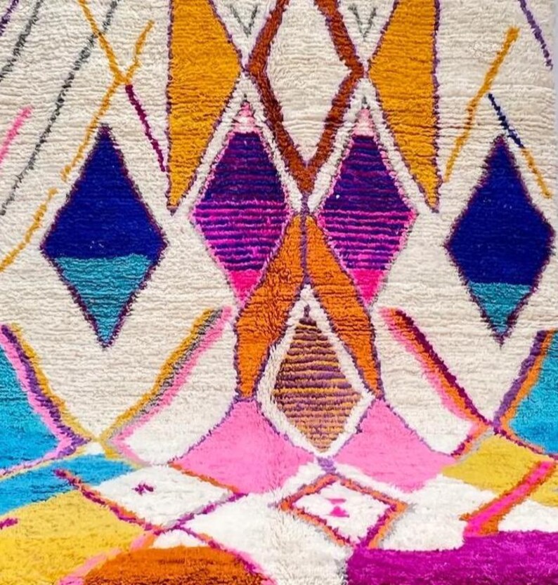 May include: Close-up of a colorful, geometric rug with a cream-colored background. The rug features diamond and triangle shapes in shades of blue, pink, orange, yellow, and purple. The rug has a textured, woven appearance.