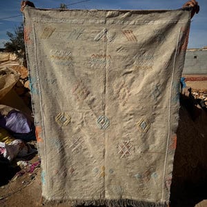 May include: Beige woven rug with geometric patterns in orange, blue, and yellow. The rug has a fringed edge and is held up against a blue sky. The rug's design includes diamond and cross shapes.