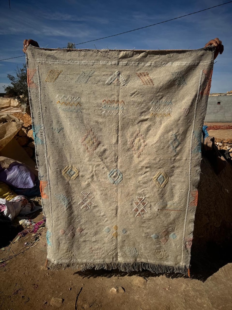 May include: Beige woven rug with geometric patterns in orange, blue, and yellow. The rug has a fringed edge and is held up against a blue sky. The rug's design includes diamond and cross shapes.