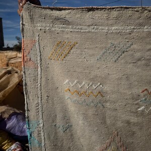 May include: A beige woven textile with colourful embroidered geometric patterns. The textile is held up against a blue sky, showcasing the intricate details of the design. The embroidery includes yellow, orange, and blue accents.