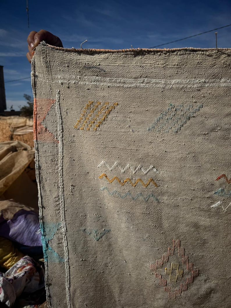 May include: A beige woven textile with colourful embroidered geometric patterns. The textile is held up against a blue sky, showcasing the intricate details of the design. The embroidery includes yellow, orange, and blue accents.