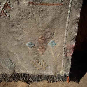 May include: A beige woven rug with geometric patterns in shades of blue, orange, and brown. The rug has a fringed edge and a textured surface, showcasing a traditional design. The rug's design includes diamond and rectangular shapes.