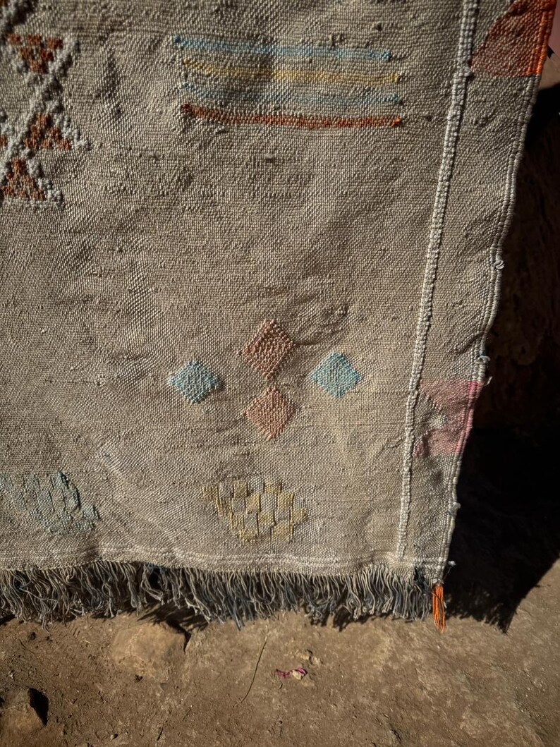 May include: A beige woven rug with geometric patterns in shades of blue, orange, and brown. The rug has a fringed edge and a textured surface, showcasing a traditional design. The rug's design includes diamond and rectangular shapes.