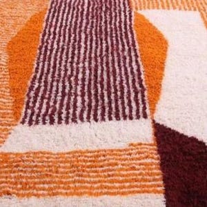 May include: Close-up of a geometric rug with a textured surface. The rug features a pattern of orange, cream, and burgundy shapes. The design includes vertical stripes and abstract forms, creating a modern aesthetic.