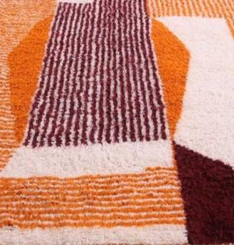 May include: Close-up of a geometric rug with a textured surface. The rug features a pattern of orange, cream, and burgundy shapes. The design includes vertical stripes and abstract forms, creating a modern aesthetic.